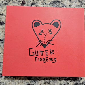 Puede incluir: Una caja roja con una ilustración negra de una cara de ratón en forma de corazón y las palabras "GUTTER FINGERS" en negro. El ratón tiene los ojos tachados y una boca cosida.
