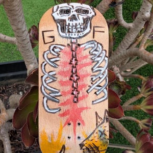 May include: A wooden skateboard deck with a hand-painted skeleton design. The artwork includes a skull, rib cage, and spine, with red and yellow accents. The letters "G" and "F" are also visible on the deck.