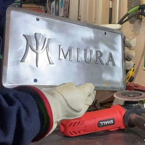 Pode incluir: Uma placa de matrícula em metal escovado com o nome "MIURA" em relevo em uma fonte serifada. A placa é segurada por uma mão enluvada, com uma ferramenta elétrica vermelha e preta visível em primeiro plano.
