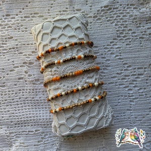 May include: Several beaded bracelets in shades of brown, orange, and cream are displayed on a white, lace-covered pillow. The bracelets are made with small, round beads and are arranged in a stacked fashion.