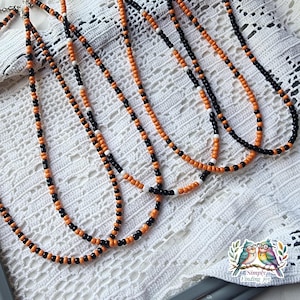 May include: Several beaded necklaces with orange, black, and white beads are displayed on a white lace surface. The necklaces have a simple, elegant design, with the beads strung together to create a long, delicate chain. The color combination is striking.