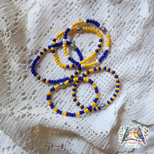 May include: Several beaded bracelets in yellow and blue, with silver-colored accents. The bracelets are arranged on a white, textured surface. The beads are small and round, creating a delicate and colorful accessory.