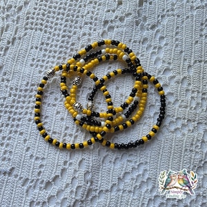 May include: A collection of beaded bracelets in yellow, black, white, and silver. The bracelets are made of small, round beads strung together. The bracelets are stacked on top of each other. The bracelets are on a white, textured surface.