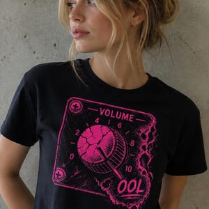 May include: Black crop top with a pink graphic of a volume knob. The graphic includes the word "VOLUME" and numbers 0-10. The word "OOL" is also visible. The shirt is worn by a person.