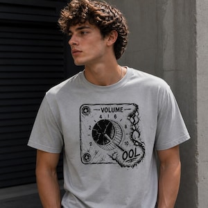 May include: A light gray t-shirt featuring a black graphic of a volume dial with the text "VOLUME" and "OOL". The design has a distressed, vintage aesthetic. The shirt is short-sleeved.