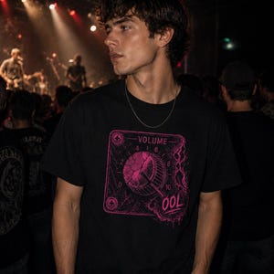 May include: Black t-shirt with a pink graphic of a volume knob and the word "OOL". The design is framed within a square with the word "VOLUME" at the top. The image is taken at a concert with a crowd and stage lights in the background.