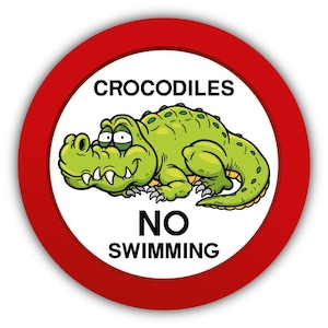 Könnte beinhalten: Rundes Schild mit rotem Rand und weißem Zentrum. Es zeigt ein Cartoon-Krokodil in Grün mit den Worten "CROCODILES" darüber und "NO SWIMMING" darunter. Das Krokodil hat weiße Zähne und lächelt.