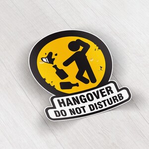 Pode incluir: Um autocolante com um círculo amarelo e uma borda preta com uma figura de pau segurando a cabeça, com garrafas e líquido derramado. O texto "HANGOVER DO NOT DISTURB" está abaixo do círculo em uma faixa branca e preta.