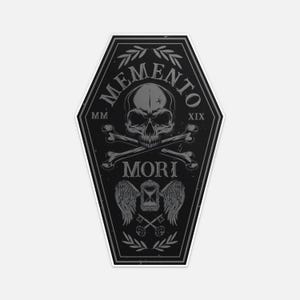 Puede incluir: Una pegatina negra con forma de ataúd con una calavera gris, huesos cruzados y las palabras "Memento Mori". El diseño incluye un reloj de arena, alas y llaves, con los números romanos MM y XIX.
