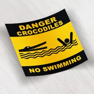 May include: A yellow and black sign with the words "DANGER CROCODILES NO SWIMMING". The sign features a black crocodile illustration and a person swimming in wavy lines, indicating a warning about crocodiles.