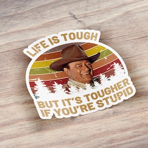 May include: A sticker featuring a portrait of a man in a cowboy hat, set against a retro sunset design with the text "LIFE IS TOUGH BUT IT'S TOUGHER IF YOU'RE STUPID". The sticker has a distressed, vintage aesthetic.
