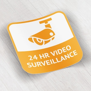 Puede incluir: Una pegatina cuadrada, naranja y blanca con esquinas redondeadas. La pegatina presenta un icono de cámara de seguridad y las palabras "24 HR VIDEO SURVEILLANCE" en texto blanco. El fondo es una veta de madera clara.