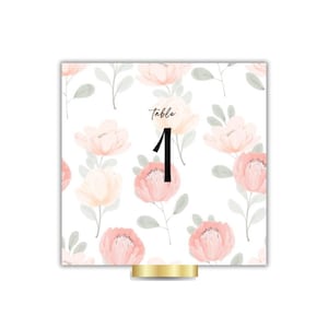 Könnte beinhalten: Eine quadratische Tischnummerkarte mit einem floralen Muster aus rosa und pfirsichfarbenen Blumen auf weißem Hintergrund. Die Zahl "1" ist in Schwarz, mit dem Wort "table" darüber. Die Karte wird von einem goldfarbenen Sockel gehalten.