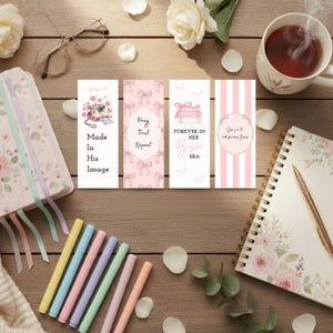 Op de afbeelding: Een verzameling briefpapierartikelen op een houten oppervlak. Bevat vier decoratieve bladwijzers met tekst, een bloemenjournaal, kleurrijke stiften, een kopje thee en een notitieboekje met een gouden pen. De bladwijzers hebben roze en witte ontwerpen.