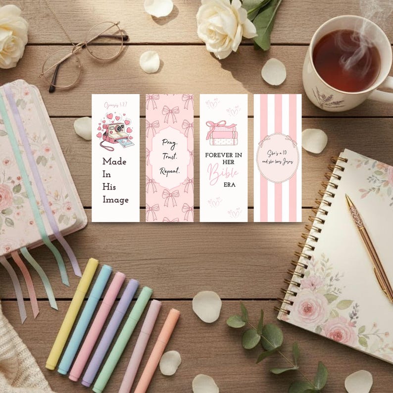 Op de afbeelding: Een verzameling briefpapierartikelen op een houten oppervlak. Bevat vier decoratieve bladwijzers met tekst, een bloemenjournaal, kleurrijke stiften, een kopje thee en een notitieboekje met een gouden pen. De bladwijzers hebben roze en witte ontwerpen.