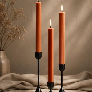 Puede incluir: Tres velas naranjas acanaladas con llamas encendidas, colocadas en candelabros de metal negro. Las velas son de diferentes alturas, creando una disposición visualmente atractiva. El fondo es neutro, resaltando las velas.