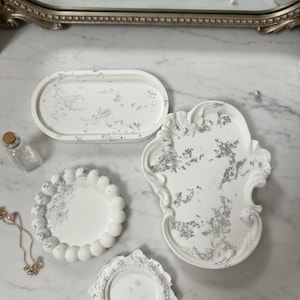 Puede incluir: Una colección de bandejas decorativas blancas en varias formas, incluyendo ovaladas, redondas y diseños ornamentados. Acentos de hojas de plata adornan las superficies. Las bandejas están dispuestas sobre una superficie de mármol, sugiriendo un tocador o exhibición.