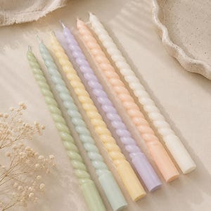 May include: A set of six long, slender taper candles with a twisted, spiral design. The candles are arranged in a row and feature a soft pastel color palette including sage green, mint, pale yellow, lavender, peach, and off-white shades.