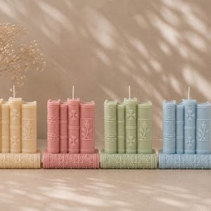 May include: Four decorative candles shaped like stacks of books. The candles are in pastel colors: cream, pink, green, and blue. Each candle has a wick and is placed on a base with a decorative pattern. The background is a neutral beige.