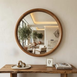 May include: A circular mirror with a wooden frame reflects an interior space. The mirror hangs above a wooden table with decorative items. The reflected space includes trees, chairs, and a reception area. The frame is made of wood.