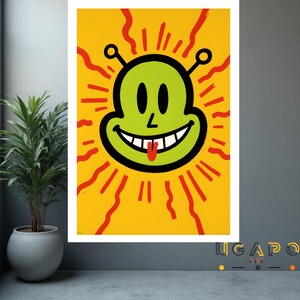 May include: A framed art print featuring a cartoon alien face with a green head, black eyes, and a red tongue sticking out. The alien is set against a yellow background with red radiating lines. The print is displayed in a room with a potted plant.