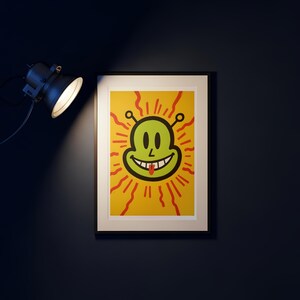 May include: Framed art print featuring a smiling alien face with a red tongue, set against a yellow background with red radiating lines. The artwork is in a black frame, illuminated by a spotlight.