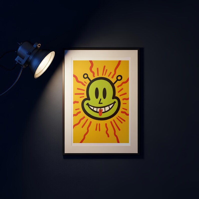 May include: Framed art print featuring a smiling alien face with a red tongue, set against a yellow background with red radiating lines. The artwork is in a black frame, illuminated by a spotlight.