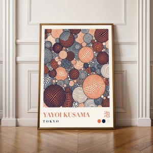 May include: A framed art print with a repeating pattern of orange, navy, and white circles. The print includes the text "YAYOI KUSAMA TOKYO". The artwork is displayed on a wooden floor. The frame is a warm, golden colour.