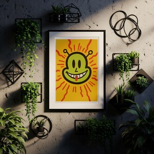 May include: A framed art print featuring a smiling green alien face with a red tongue and radiating lines on a yellow background. The artwork is in a black frame and is displayed on a textured gray wall, surrounded by decorative plants.