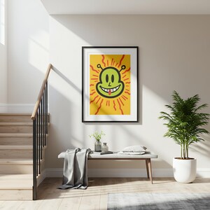 May include: A framed art print featuring a cartoon alien face with a smiling expression and a red tongue. The alien is green with black eyes and antennae, set against a yellow background with red sun rays. The artwork is displayed in a black frame.