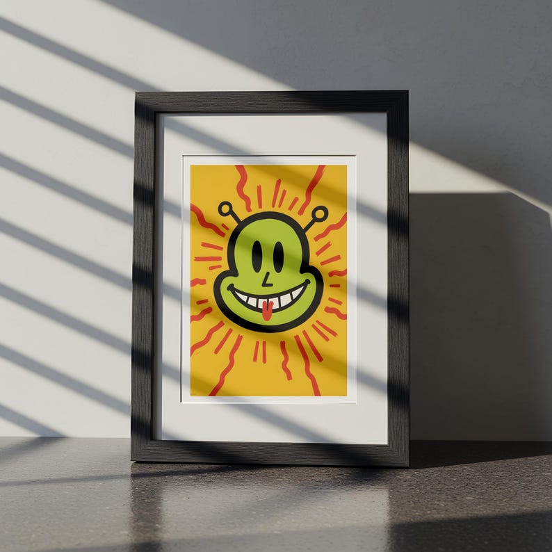 May include: Framed art print featuring a smiling alien face with a protruding tongue, set against a yellow background with red radiating lines. The alien is green with black details. The artwork is displayed in a black frame.