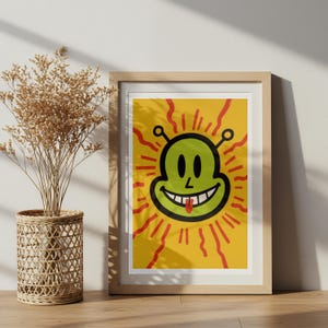 May include: A framed art print featuring a smiling green alien face with a red tongue and antenna on a yellow background. The artwork has red sunburst rays emanating from the alien's head. The frame is made of light-colored wood.