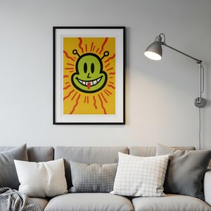 May include: A framed art print featuring a smiling alien face with a red tongue, set against a yellow background with red rays. The alien is green with black outlines. The artwork is displayed above a gray sofa with pillows.