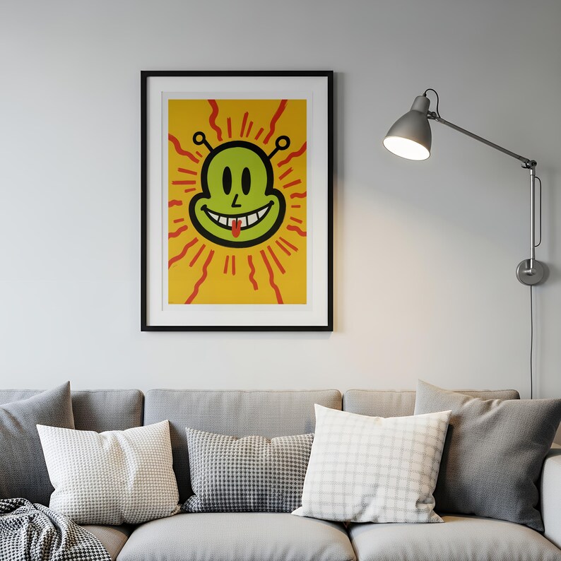 May include: A framed art print featuring a smiling alien face with a red tongue, set against a yellow background with red rays. The alien is green with black outlines. The artwork is displayed above a gray sofa with pillows.