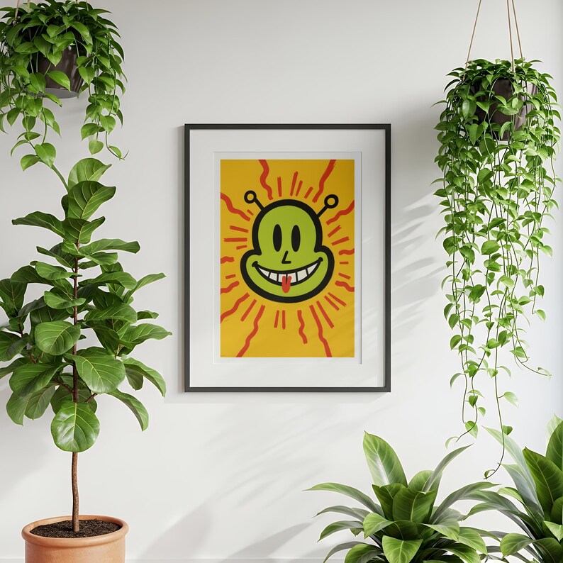 May include: A framed art print featuring a smiling alien face with a red tongue, set against a yellow background with red sun rays. The alien is green with black outlines. The artwork is in a black frame.