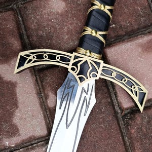 May include: A close-up of a decorative sword with a silver blade and a black handle wrapped in leather. The hilt features gold accents and a stylized design. The sword is set against a brick background.