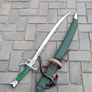 Puede incluir: Una espada de fantas&iacute;a con una hoja plateada y un mango verde, junto con su vaina verde. La vaina tiene correas de cuero marr&oacute;n. La espada y la vaina se exhiben sobre una superficie pavimentada gris.