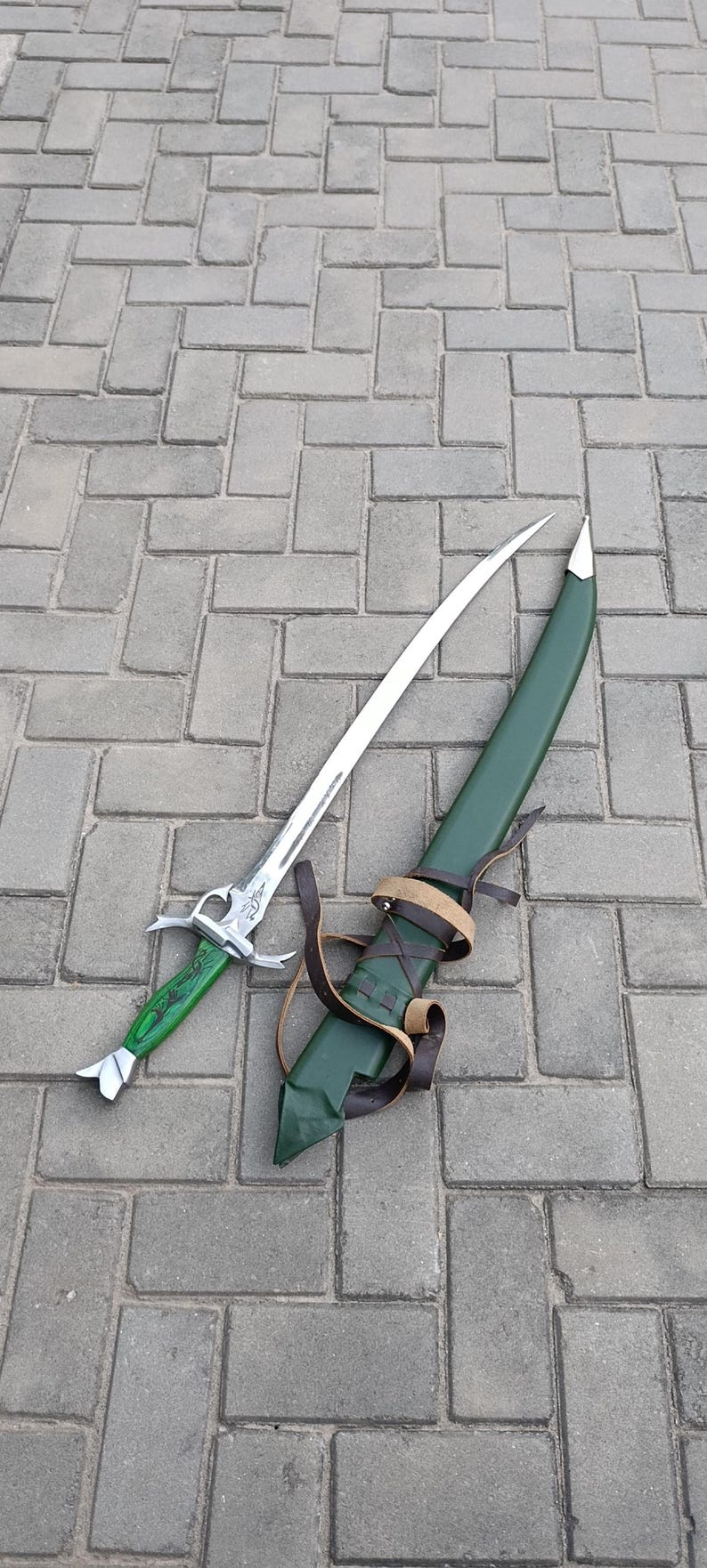 Puede incluir: Una espada de fantas&iacute;a con una hoja plateada y un mango verde, junto con su vaina verde. La vaina tiene correas de cuero marr&oacute;n. La espada y la vaina se exhiben sobre una superficie pavimentada gris.