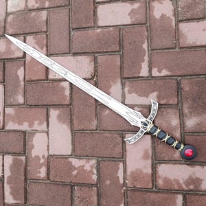 May include: A decorative sword with a silver blade and intricate designs. The hilt is black with gold accents and a red gem at the pommel. The sword is lying on a brick surface.