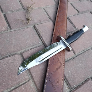 May include: A hunting knife with a polished steel blade and a black handle rests on a brown leather sheath. The knife has a silver bolster and a curved blade. The sheath is embossed with a decorative pattern.