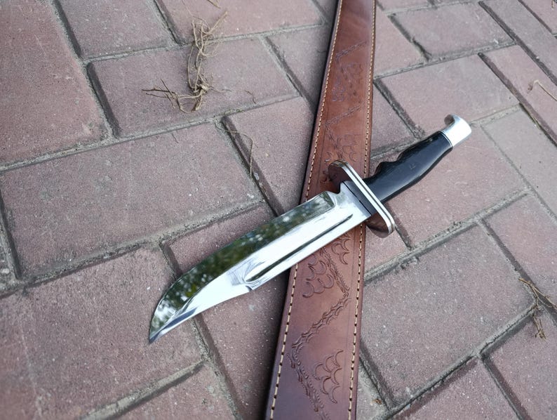 May include: A hunting knife with a polished steel blade and a black handle rests on a brown leather sheath. The knife has a silver bolster and a curved blade. The sheath is embossed with a decorative pattern.