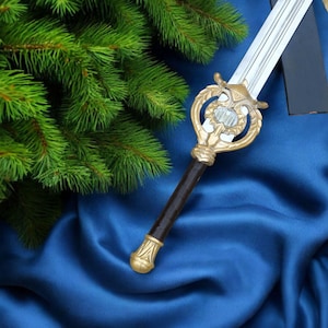 Puede incluir: Una espada decorativa con empuñadura dorada y mango marrón oscuro, sobre un fondo de tela azul. La hoja de la espada es plateada. Ramas verdes de pino en el fondo.