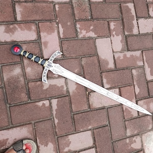 May include: A decorative sword with a silver blade and a black and gold handle. The sword features a red gem at the pommel and a crossguard design. The sword is lying on a brick surface.