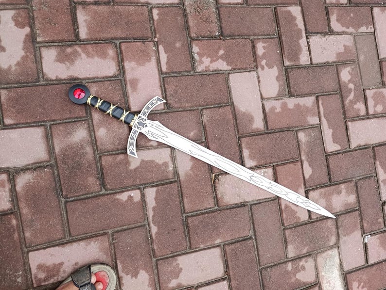 May include: A decorative sword with a silver blade and a black and gold handle. The sword features a red gem at the pommel and a crossguard design. The sword is lying on a brick surface.