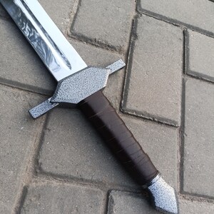 May include: A silver-bladed sword with a textured gray crossguard and pommel. The handle is wrapped in dark brown leather. The sword is displayed on a gray brick surface.