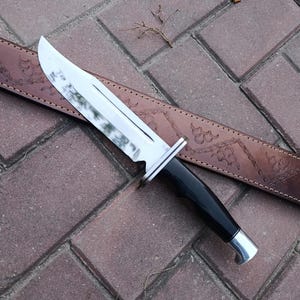 May include: A large hunting knife with a shiny, silver blade and a black handle. The knife rests on a brown leather sheath with decorative embossed patterns. The knife is designed for outdoor use.