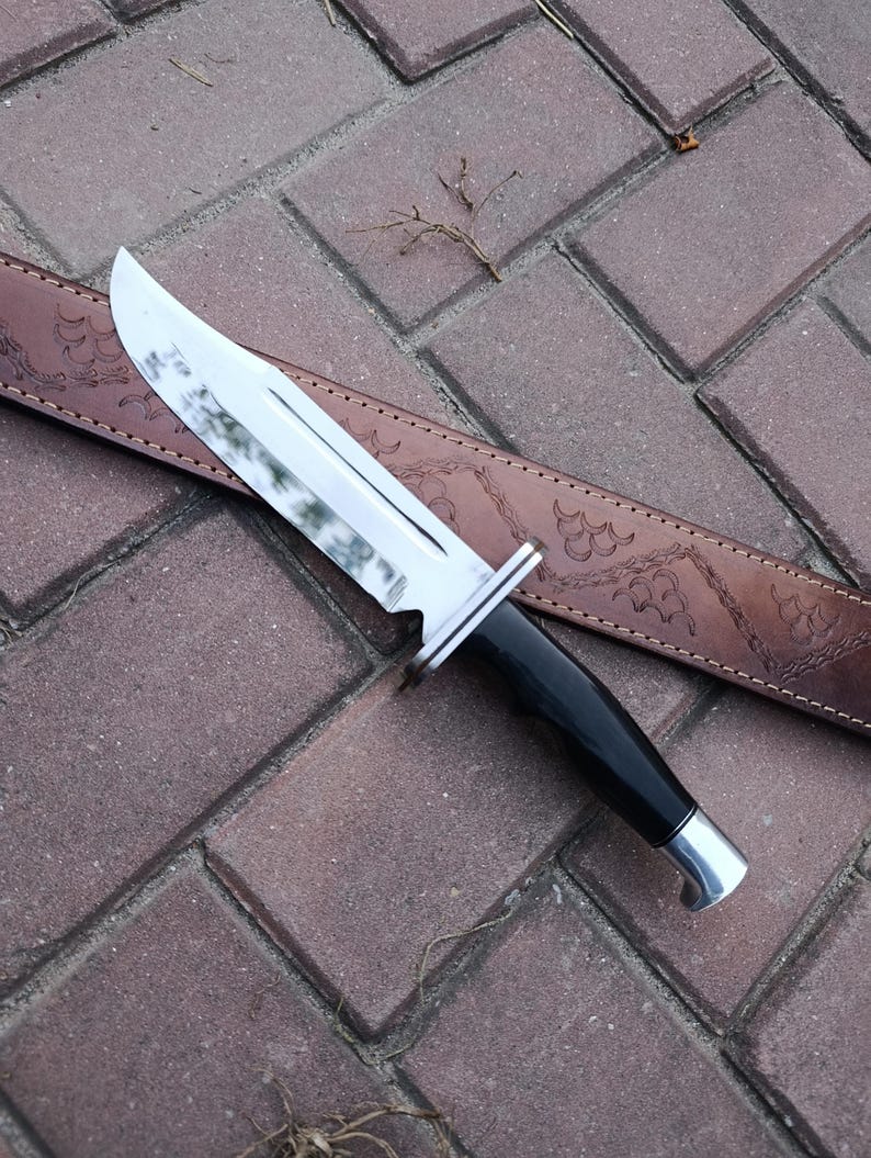 May include: A large hunting knife with a shiny, silver blade and a black handle. The knife rests on a brown leather sheath with decorative embossed patterns. The knife is designed for outdoor use.