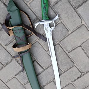 Puede incluir: Una espada de estilo fantas&iacute;a con una hoja curva y un mango verde con tallas decorativas. La espada est&aacute; junto a una vaina verde con correas de cuero marr&oacute;n. La hoja mide aproximadamente 46 cm de largo.