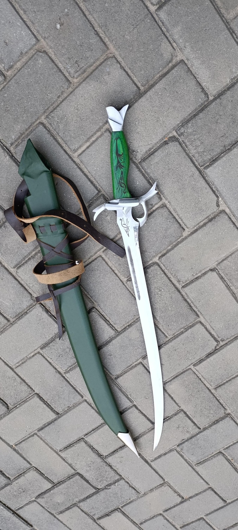 Puede incluir: Una espada de estilo fantas&iacute;a con una hoja curva y un mango verde con tallas decorativas. La espada est&aacute; junto a una vaina verde con correas de cuero marr&oacute;n. La hoja mide aproximadamente 46 cm de largo.