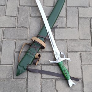 Puede incluir: Una espada plateada con una hoja curva y un mango verde, apoyada sobre una vaina verde. La espada tiene un dise&ntilde;o ornamentado en el mango y una correa de cuero marr&oacute;n.